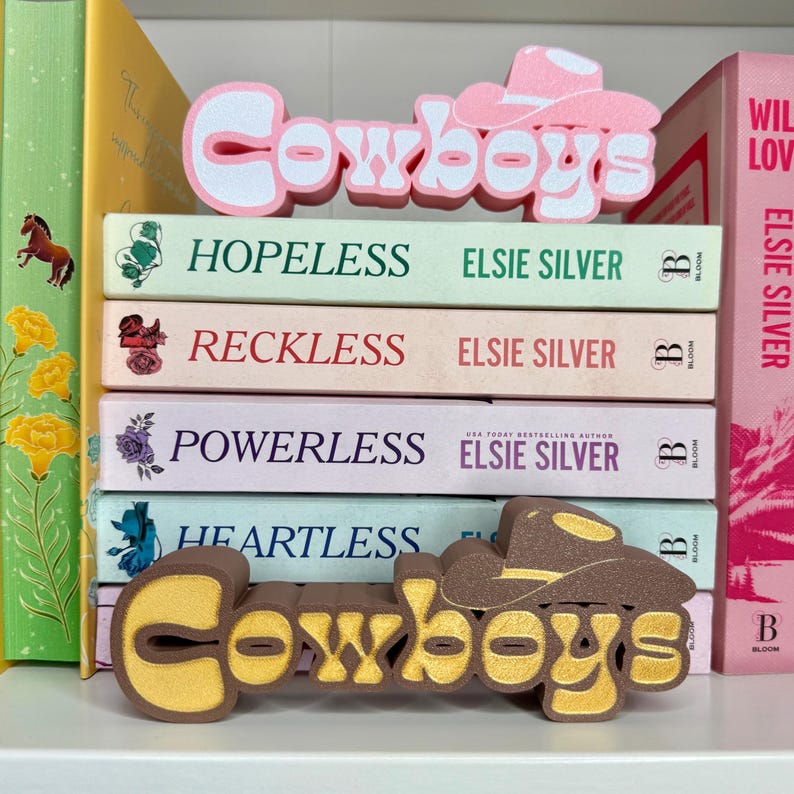 Puede incluir: Una pila de libros con t&iacute;tulos como "Hopeless", "Reckless", "Powerless" y "Heartless" de Elsie Silver. Los libros est&aacute;n decorados con sujetalibros con tem&aacute;tica de vaqueros en rosa y marr&oacute;n, con la palabra "Cowboys".