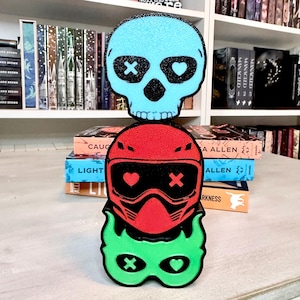 May include: A stack of three colorful, glittery cutouts: a light blue skull with black cross and heart eyes, a red helmet with black visor and heart and cross details, and a green mask with heart and cross eyes. The cutouts are stacked on a white surface.