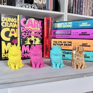 May include: Four colorful cat figurines wearing crowns and sunglasses are displayed in front of a bookshelf with books. The cats are yellow, pink, blue, and gold. The books have titles like "Dung Craw" and "Carl Doomsday Scenario."