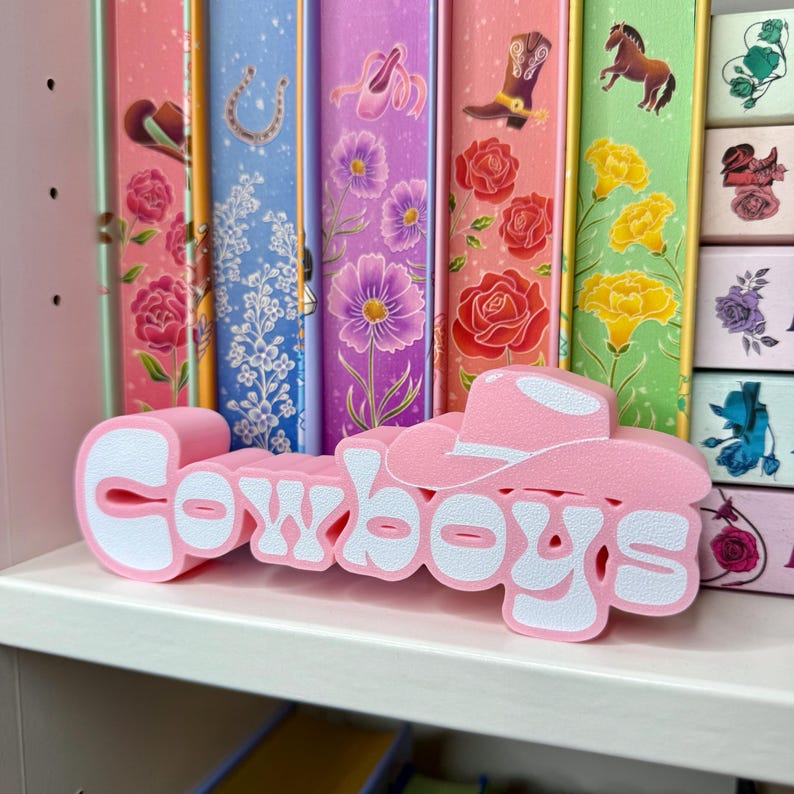 Puede incluir: Un letrero decorativo rosa y blanco que dice "Cowboys" en una fuente juguetona, coronado con un sombrero de vaquero. El letrero est&aacute; sobre un estante blanco, con coloridos sujetalibros con temas florales y del oeste en el fondo.