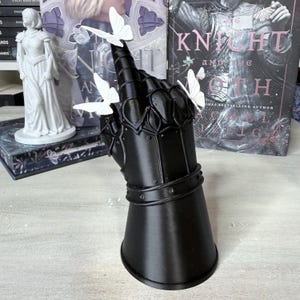 May include: A black gauntlet sculpture shaped like a hand with fingers pointing upward, featuring articulated metal-like plating details. Three small, white, three-dimensional butterflies are attached to the fingers and thumb of the gauntlet.