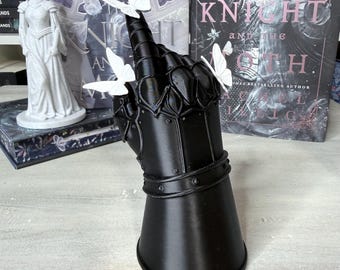 May include: A black gauntlet sculpture shaped like a hand with fingers pointing upward, featuring articulated metal-like plating details. Three small, white, three-dimensional butterflies are attached to the fingers and thumb of the gauntlet.