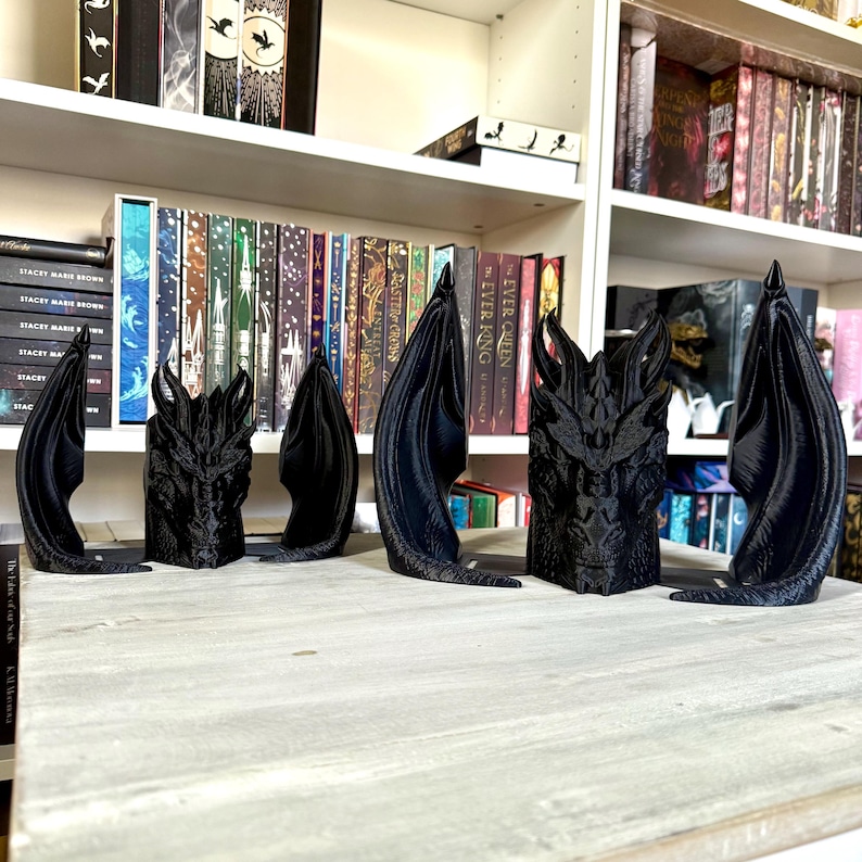 May include: Four black dragon bookends with wings spread out. The bookends are on a wooden surface in front of a bookshelf filled with books.