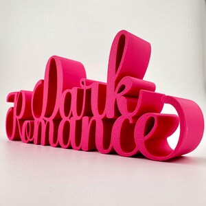 Puede incluir: Un letrero rosa impreso en 3D que dice "Dark Romance". Las letras son estilizadas y tienen una superficie texturizada.