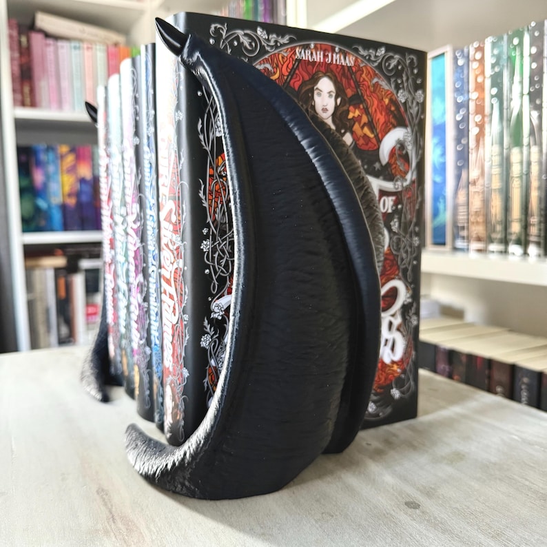 May include: Black bookends shaped like a pair of demon wings holding up a stack of books. The bookends are made of a smooth, matte black plastic. The bookends are holding up a stack of books, including one with the title "Queen of the Conquered".
