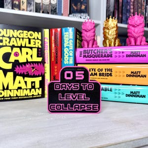 May include: A black and pink sign with the text "05 DAYS TO LEVEL COLLAPSE" stands in front of a stack of colorful books. The books have titles like "Dungeon Crawler Carl" and "The Butcher's Masquerade". Pink and gold cat figurines sit on top of the books.