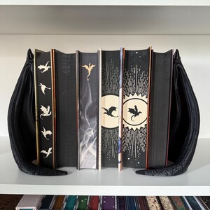 May include: A set of seven black hardcover books with dragon silhouettes on the spines. The books are arranged on a white shelf with black bookends shaped like dragon wings.