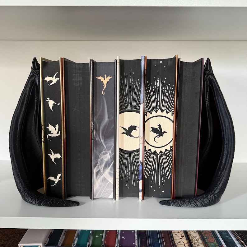 May include: A set of seven black hardcover books with dragon silhouettes on the spines. The books are arranged on a white shelf with black bookends shaped like dragon wings.