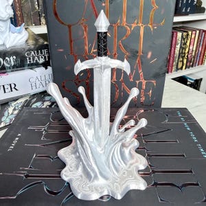 May include: A silver sword sculpture with a black handle, set in a splash-like base. The sculpture is in front of a book with the title "Callie Hart" and "Stone". The background includes other books.
