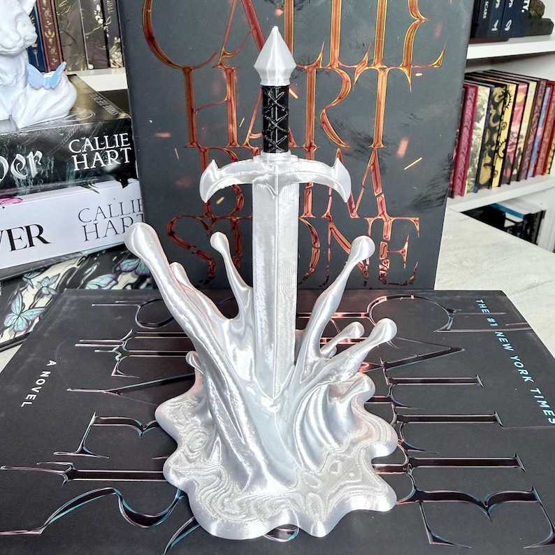 May include: A silver sword sculpture with a black handle, set in a splash-like base. The sculpture is in front of a book with the title "Callie Hart" and "Stone". The background includes other books.