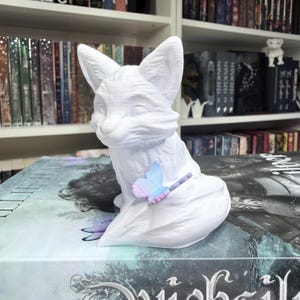 May include: A white fox figurine with large ears and a seated pose. A small, colorful butterfly rests on its tail. The fox is placed on top of a book with a dark cover. The background includes a bookshelf filled with books.