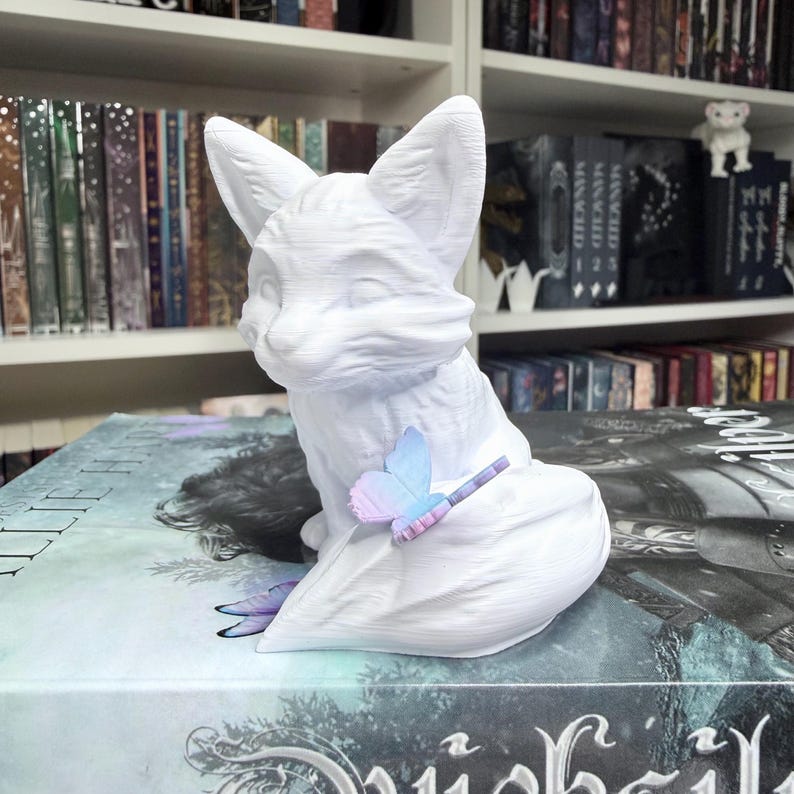 May include: A white fox figurine with large ears and a seated pose. A small, colorful butterfly rests on its tail. The fox is placed on top of a book with a dark cover. The background includes a bookshelf filled with books.