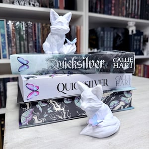 May include: Two white fox figurines sit atop a stack of books. The books are titled "Quicksilver" by Callie Hart, with butterfly illustrations. The figurines are detailed and the butterflies are in shades of blue, pink, and purple.