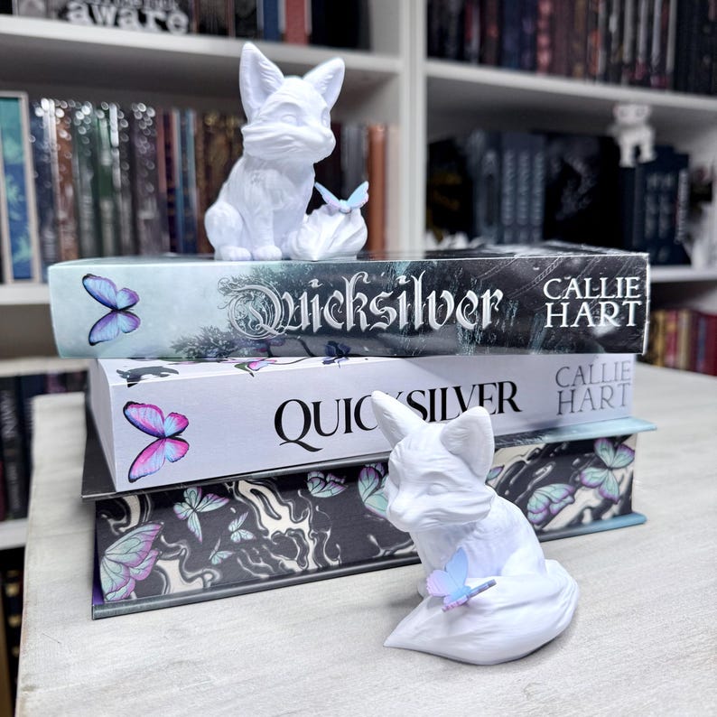 May include: Two white fox figurines sit atop a stack of books. The books are titled "Quicksilver" by Callie Hart, with butterfly illustrations. The figurines are detailed and the butterflies are in shades of blue, pink, and purple.