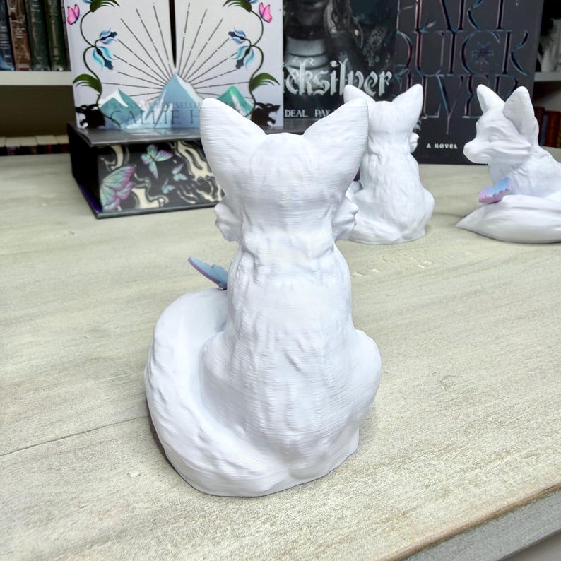 May include: White fox figurines with a sitting pose and textured fur. One fox has a blue butterfly accent. Other decorative items are visible in the background, including books and a small box.