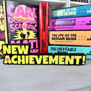 May include: A stack of books with colorful covers, including "Carl's Doomsday Scenario" and "Dungeon Anarchist's Cookbook." A black and yellow sign reads "NEW ACHIEVEMENT!" in bold letters. The books are on a white surface.