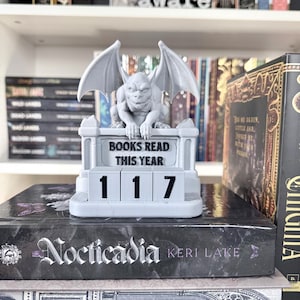 May include: A gray gargoyle book tracker with the text "BOOKS READ THIS YEAR" and the number "117". The gargoyle sits atop a book titled "Nocticaida" by Keri Lake. Bookshelves are in the background.