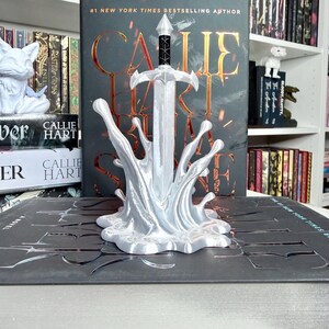 May include: A silver-colored sword bookend with a splash design, standing in front of a book. The book has the title "CALLIE HART" and is by a #1 New York Times Bestselling Author. Other books are visible in the background.