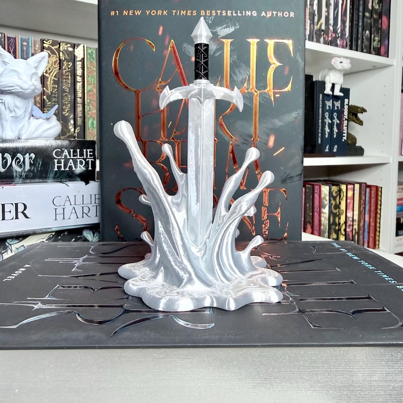 May include: A silver-colored sword bookend with a splash design, standing in front of a book. The book has the title "CALLIE HART" and is by a #1 New York Times Bestselling Author. Other books are visible in the background.
