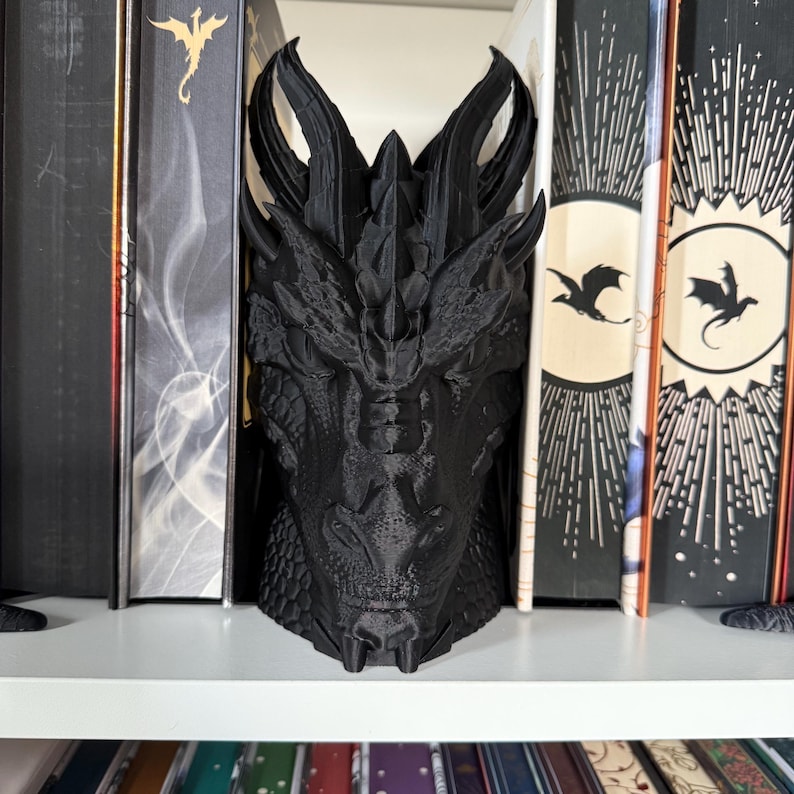 May include: Black dragon head bookends. The dragon is detailed with scales and has a fierce expression. The bookends are sitting on a white bookshelf.