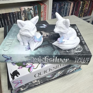 May include: Two white fox figurines with butterfly accents rest on top of books. The books are titled "Quicksilver" by Callie Hart. The background includes a bookshelf filled with books.
