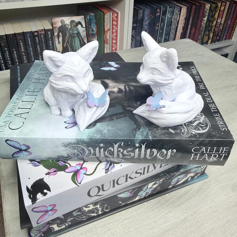 May include: Two white fox figurines with butterfly accents rest on top of books. The books are titled "Quicksilver" by Callie Hart. The background includes a bookshelf filled with books.