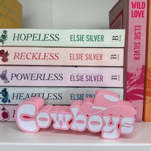 Puede incluir: Un objeto decorativo rosa y blanco con tem&aacute;tica de Cowboys, con la forma de la palabra "Cowboys" con un dise&ntilde;o de sombrero de vaquero. Se coloca delante de una pila de libros con t&iacute;tulos como "Hopeless" y "Reckless" de Elsie Silver.