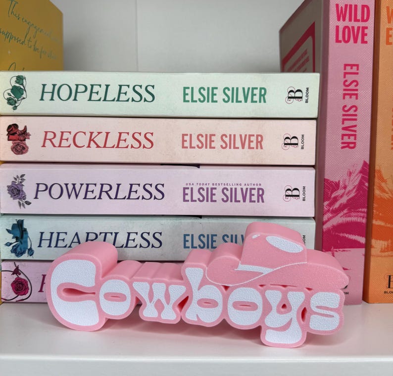 Puede incluir: Un objeto decorativo rosa y blanco con tem&aacute;tica de Cowboys, con la forma de la palabra "Cowboys" con un dise&ntilde;o de sombrero de vaquero. Se coloca delante de una pila de libros con t&iacute;tulos como "Hopeless" y "Reckless" de Elsie Silver.