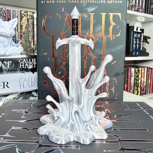 May include: A silver-colored sword sculpture with a splash design base. The sword has a black handle and a pointed tip. The background features books and the text "CALLIE HART".