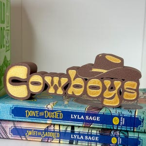 Puede incluir: Un letrero decorativo marr&oacute;n y dorado que dice "Cowboys" con un dise&ntilde;o de sombrero de vaquero. El letrero est&aacute; colocado encima de dos libros con cubiertas azules. Los libros se titulan "Done and Dusted" y "Swift and Saddled" de Lyla Sage.