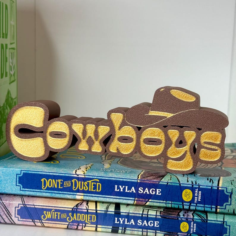 Puede incluir: Un letrero decorativo marr&oacute;n y dorado que dice "Cowboys" con un dise&ntilde;o de sombrero de vaquero. El letrero est&aacute; colocado encima de dos libros con cubiertas azules. Los libros se titulan "Done and Dusted" y "Swift and Saddled" de Lyla Sage.