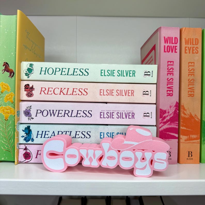 Puede incluir: Una colecci&oacute;n de libros de Elsie Silver, incluyendo "Hopeless" y "Reckless", con un recorte rosa y blanco de "Cowboys". Los libros est&aacute;n apilados en una estanter&iacute;a blanca, con otros libros en verde, amarillo, rosa y naranja.