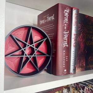 May include: A red and black 3D printed seven-pointed star bookend. The bookend is shaped like a circle and has a raised design. The bookend is sitting on a white shelf with two books behind it. The books are titled "Empire of the Vampire" and "Empire of the Damned".