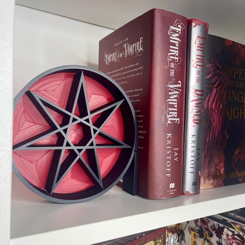 May include: A red and black 3D printed seven-pointed star bookend. The bookend is shaped like a circle and has a raised design. The bookend is sitting on a white shelf with two books behind it. The books are titled "Empire of the Vampire" and "Empire of the Damned".