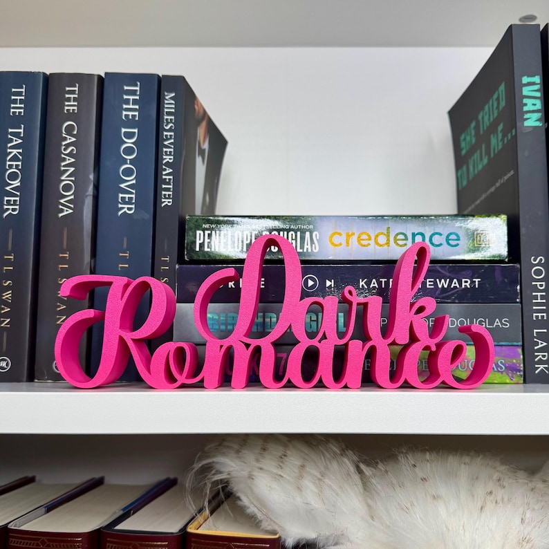 Puede incluir: Un letrero de madera rosa con las palabras "Dark Romance" en una fuente estilizada. El letrero est&aacute; sentado en una estanter&iacute;a con varios libros detr&aacute;s.