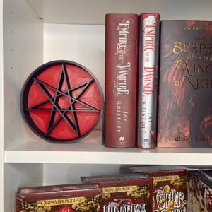 May include: A red and black 3D printed seven-pointed star, also known as a heptagram, on a white shelf with books behind it. The books are titled "Empire of the Vampire" and "Empire of the Damned" by Jay Kristoff, and "The Serpent and the Wings of Night" by Carissa Broadbent.