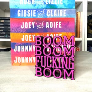 May include: A stack of colorful books with names on the spines, including "Hugh and Lizzie," "Gibsie and Claire," and "Joey and Aoife." In front, a pink and black sign reads "BOOM BOOM FUCKING BOOM."