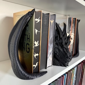 May include: A pair of black dragon bookends holding up a row of books. The bookends are detailed and have a textured finish. The book on the left has the title "The Lord of the Rings" on the spine.