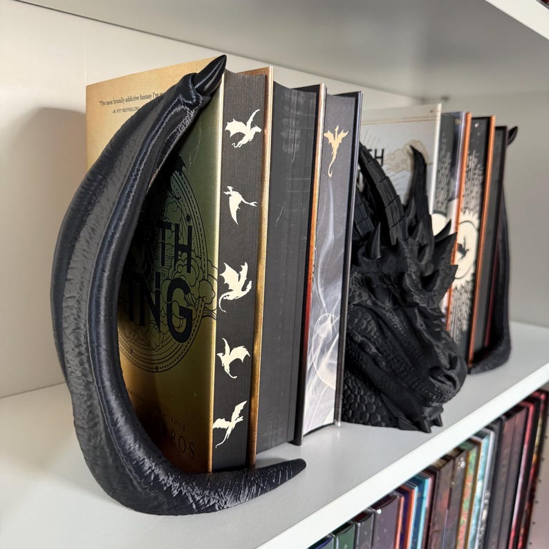 May include: A pair of black dragon bookends holding up a row of books. The bookends are detailed and have a textured finish. The book on the left has the title "The Lord of the Rings" on the spine.