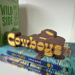 Puede incluir: Una pieza decorativa marr&oacute;n y dorada con la palabra "Cowboys" y un dise&ntilde;o de sombrero de vaquero. Se coloca sobre dos libros titulados "Done and Dusted" y "Swift and Saddled" de Lyla Sage. Un libro verde titulado "Wild Side" se ve en el fondo.