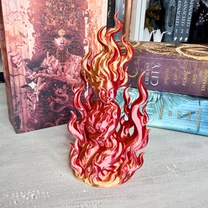 May include: A red and gold figurine of a woman engulfed in flames. The sculpture has a detailed design with flowing hair and flames. The figurine is likely a decorative item or a piece of art. The color gradient goes from gold to red.