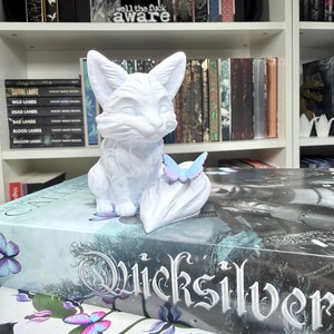 May include: A white fox figurine with a butterfly perched on a leaf-shaped base. The fox is sitting upright with its ears perked. The figurine is on top of a book with the title "Quicksilver". The text "well the fuck aware" is visible on a shelf in the background.