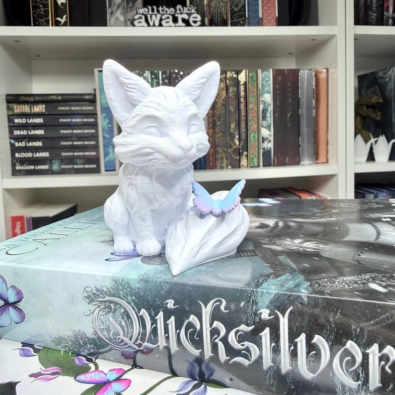 May include: A white fox figurine with a butterfly perched on a leaf-shaped base. The fox is sitting upright with its ears perked. The figurine is on top of a book with the title "Quicksilver". The text "well the fuck aware" is visible on a shelf in the background.
