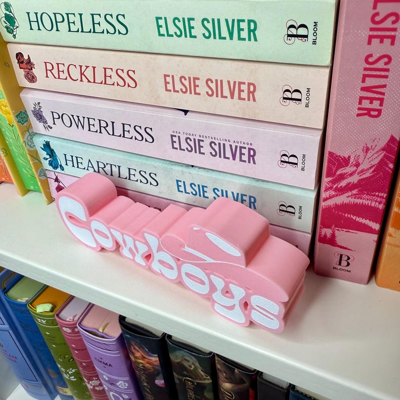 Puede incluir: Un adorno rosa para estanter&iacute;a con la palabra "Cowboys" en letras blancas. Se encuentra frente a una pila de libros con t&iacute;tulos como "Hopeless" y "Reckless" de Elsie Silver, creando una composici&oacute;n visual.