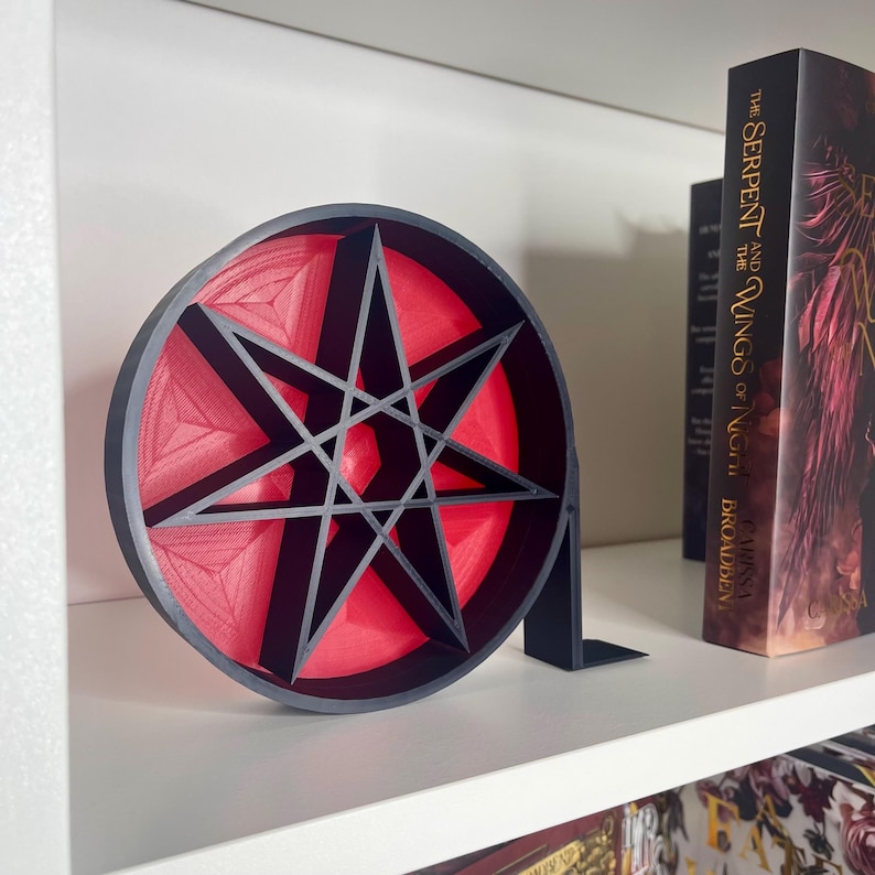 May include: A 3D printed black and red bookend shaped like a seven-pointed star. The star is hollow and has a red interior. The bookend is on a white shelf with a book titled "The Serpent and the Wings of Night" by Laini Taylor.