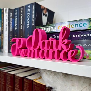 Puede incluir: Un letrero de madera rosa con las palabras "Dark Romance" en una fuente estilizada. El letrero est&aacute; sentado en una estanter&iacute;a con otros libros.