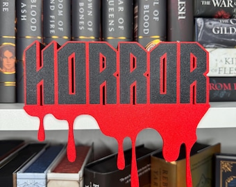 May include: A black and red wooden bookend shaped like the word "HORROR" with dripping blood. The bookend is sitting on a shelf with books behind it.