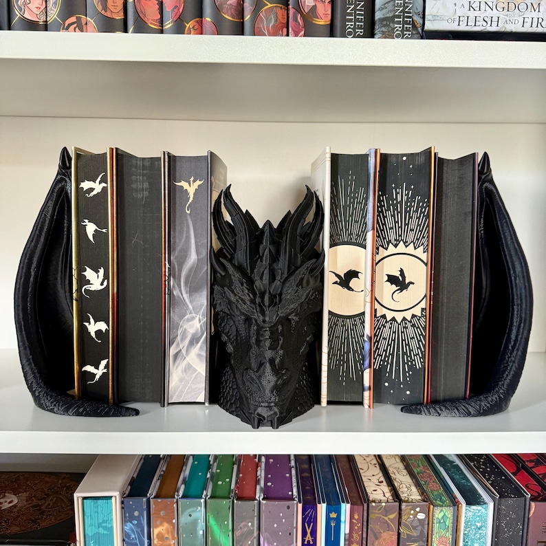 May include: Black dragon bookends with a 3D dragon head in the center. The bookends are holding up a row of books with black covers and white designs. The books on the bottom shelf have colorful covers.