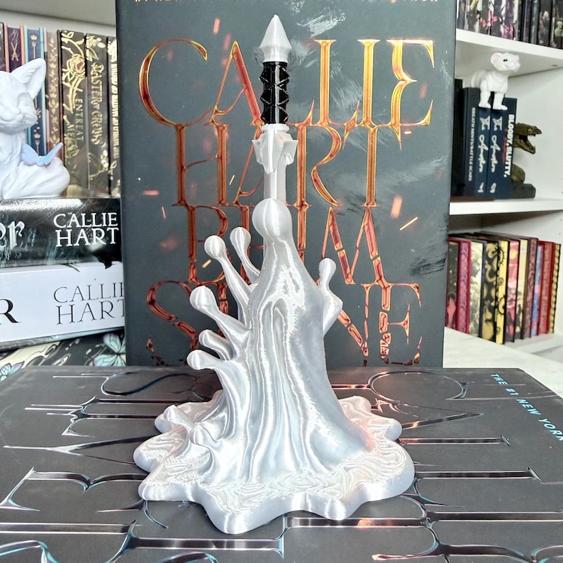 May include: A 3D-printed, silver-colored book stand shaped like a stylized splash. The stand holds a book with the title "CALLIE HART" visible. The stand has a black and white decorative element at the top. Other books are visible in the background.