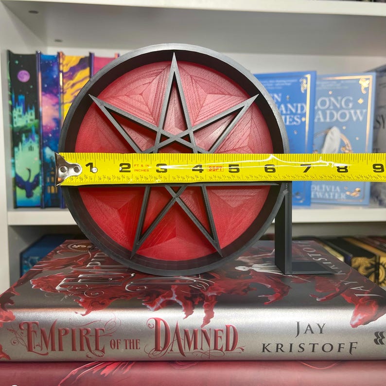 May include: A black and red 3D printed star with six points, measuring 6 inches in diameter, is displayed on a shelf with books. The star is on a black stand.